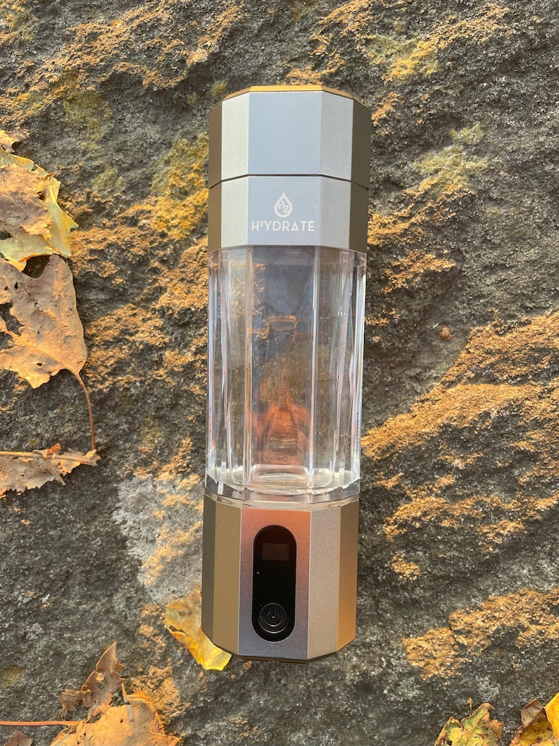 May include: A gray and silver water bottle purifier with a hexagonal design. The bottle has a clear, cylindrical reservoir and a dark gray base. The brand name 'Hydrate' is subtly printed on the bottle. The device appears to be portable and suitable for outdoor use.