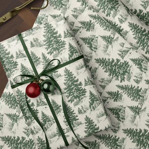 May include: Gift wrapping paper with a green and white forest pattern. A wrapped gift is adorned with a green ribbon and two ornaments. A roll of paper, scissors, and tree-shaped paper clips are also visible.