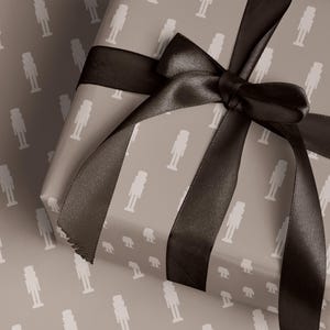 Könnte beinhalten: Ein verpacktes Geschenk mit grauem Hintergrund und weißem Nussknackermuster. Das Geschenk ist mit einem dunkelbraunen Satinband und einer Schleife gebunden. Geschenkpapier und Band erzeugen ein festliches Urlaubsgefühl.