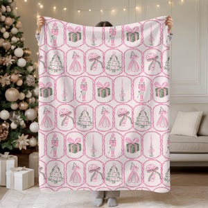 May include: A pink and white Christmas-themed blanket featuring a repeating pattern of holiday icons. The design includes nutcrackers, wrapped gifts, Christmas trees, ballerinas, and candy canes, all framed in decorative ovals.