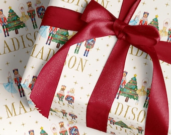 May include: Cream-coloured gift wrap with a repeating pattern of nutcrackers, Christmas trees, and ballerinas. The name "MADISON" is printed in gold. A burgundy ribbon is tied in a bow around the wrapped gift.