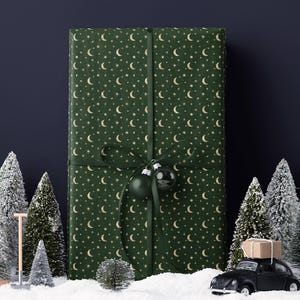 May include: A large gift wrapped in green paper with a pattern of gold moons and stars, tied with a green ribbon and adorned with two green ornaments. The scene includes miniature snowy trees and a black toy car carrying a package.
