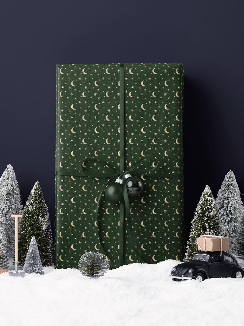 May include: A large gift wrapped in green paper with a pattern of gold moons and stars, tied with a green ribbon and adorned with two green ornaments. The scene includes miniature snowy trees and a black toy car carrying a package.