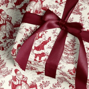 Puede incluir: Un regalo de Navidad envuelto en papel con un diseño toile de Jouy rojo y blanco con motivos navideños. Una cinta de raso burdeos está atada en un lazo en la parte superior. El regalo está listo para ser entregado.