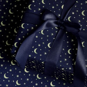May include: A wrapped gift with navy blue paper featuring a pattern of gold crescent moons and stars. A matching navy blue satin ribbon is tied in a bow on top, creating a festive and celestial theme.