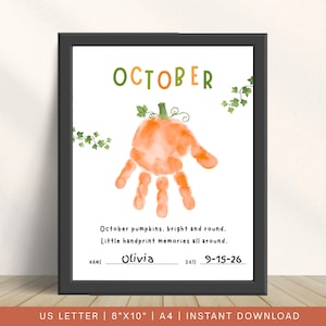 May include: A framed print featuring the word OCTOBER in green and orange, a central orange handprint shaped like a pumpkin with green vines, and the text: October pumpkins, bright and round. Little handprint memories all around. NAME Olivia DATE 9-15-26.