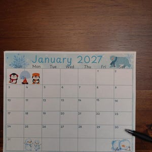 May include: A January 2027 calendar page featuring a light blue border with snowflake patterns. The grid includes illustrations of penguins, an igloo, a campfire, snowmen, and a polar bear. A black pen rests on the bottom right corner.