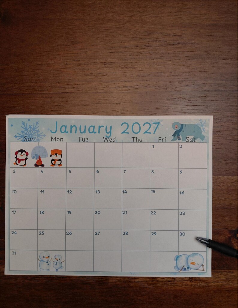 May include: A January 2027 calendar page featuring a light blue border with snowflake patterns. The grid includes illustrations of penguins, an igloo, a campfire, snowmen, and a polar bear. A black pen rests on the bottom right corner.