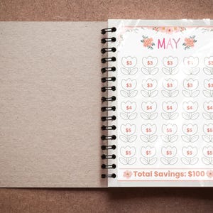 May include: A spiral-bound notebook open to a savings tracker page for May. The page features a floral design with tulip-shaped boxes, each with a dollar amount. The total savings goal is $100.