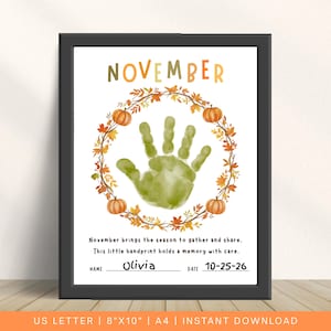 May include: A framed print featuring the word NOVEMBER in multicolored letters above a wreath of autumn leaves and pumpkins. A green handprint sits in the center. Text reads: November brings the season to gather and share. This little handprint holds a memory with care. Name: Olivia, Date: 10-25-26.