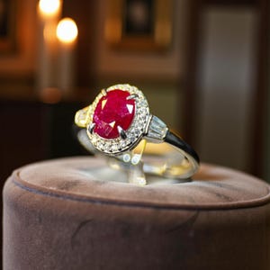 May include: A ruby ring is displayed. The ring has a large, oval-cut ruby, encircled by small diamonds. The band is silver with black detailing and baguette diamonds on either side of the ruby, showcasing a classic design.