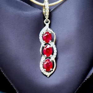 May include: A gold pendant necklace featuring three oval, vibrant red gemstones. Each gemstone is set within a decorative, openwork frame, embellished with small, sparkling clear stones. The pendant hangs from a delicate gold chain.