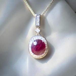 May include: A pendant necklace featuring a large, oval-cut deep red gemstone surrounded by a halo of small, clear baguette-cut stones. The pendant hangs from a delicate silver-toned chain and includes a rectangular and square stone accent.