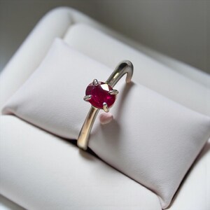 May include: A silver ring with a vibrant red oval gemstone. The ring's band is a light gold color, curving upwards to cradle the gemstone. The ring is displayed on a white, textured surface.