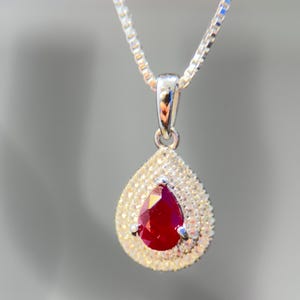 May include: A silver necklace with a teardrop pendant. The pendant features a vibrant red gemstone surrounded by a halo of small, sparkling stones. The chain is a delicate box chain style.