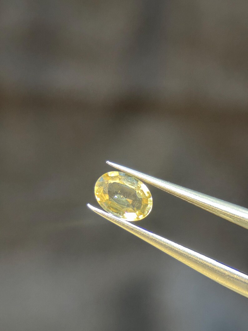 May include: A yellow oval gemstone held by silver tweezers. The gemstone has a light yellow colour and a polished surface. The tweezers are angled, showcasing the gemstone against a blurred background. The gemstone is likely a sapphire or topaz.