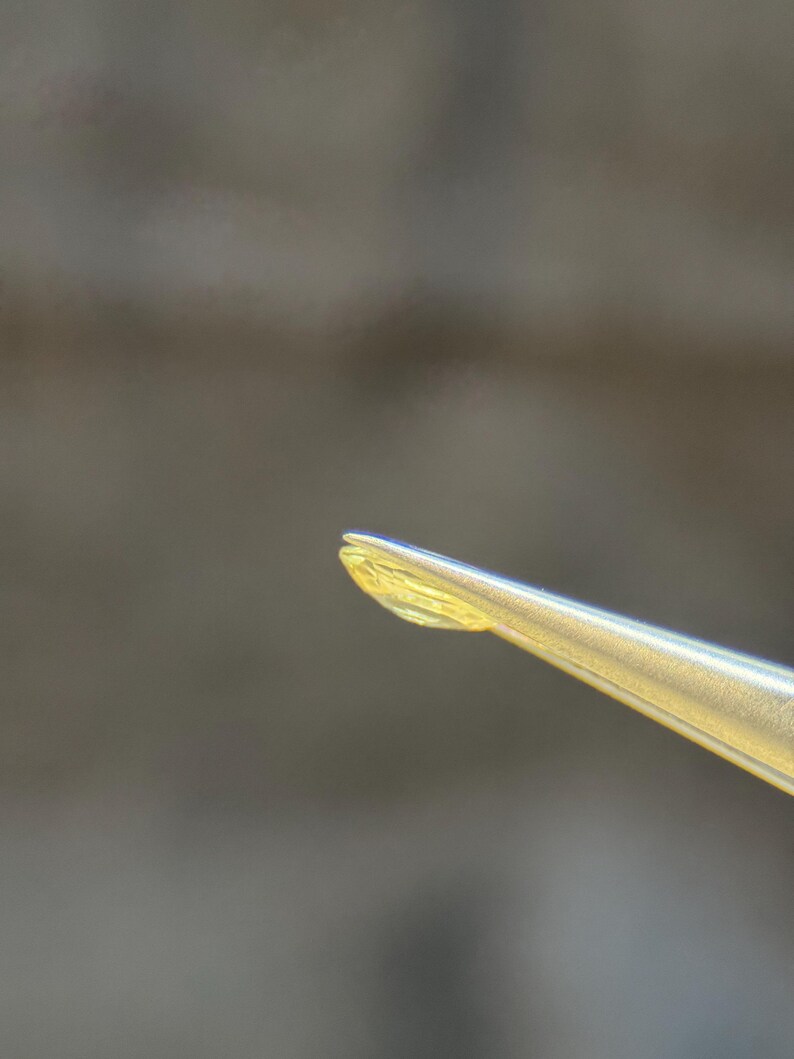 May include: Close-up of a gold-coloured tool, possibly tweezers, holding a small, translucent, yellow object. The object has a slightly curved shape. The background is blurred, with shades of brown and grey.