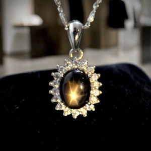 May include: A silver pendant necklace featuring a black star sapphire gemstone. The oval-shaped gemstone has a star-like pattern and is surrounded by a halo of small, clear gemstones. The necklace chain is also silver.