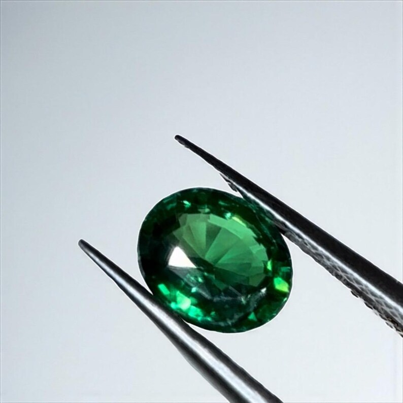 May include: A faceted, oval-cut, emerald-green gemstone is held by a pair of silver tweezers against a white background. The gemstone reflects light, showcasing its clarity and brilliance. The tweezers are positioned to display the gem.