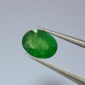 May include: A vibrant, oval-cut emerald gemstone, held by silver tweezers. The gem displays a rich, deep green color with visible facets. The tweezers are positioned to showcase the gemstone against a neutral background, highlighting its clarity and brilliance.