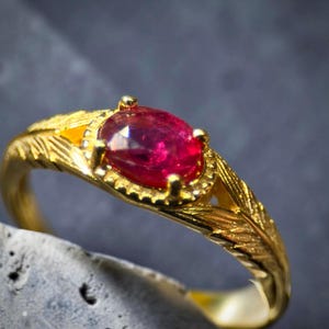 May include: A gold ring featuring a central oval-cut deep red ruby gemstone. The band is crafted with a textured, leaf-like pattern that extends from the setting. The ruby is held in place by four prongs within a decorative, scalloped metal border.