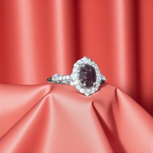 May include: A silver ring with a large, oval, dark purple gemstone at the center, surrounded by a halo of smaller, clear stones. The ring is set against a red fabric background.