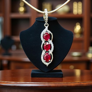 May include: A silver pendant necklace with three oval, deep red gemstones. The gemstones are surrounded by a halo of small, clear crystals. The pendant hangs from a delicate gold chain and is displayed on a black jewelry stand.