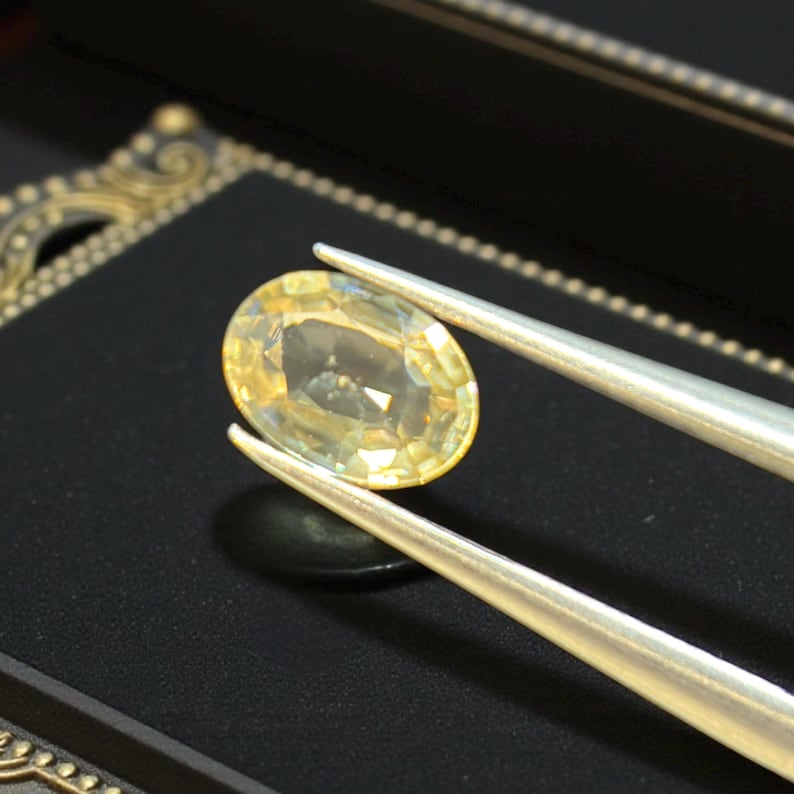May include: A pale yellow, oval gemstone is held by silver tweezers against a black backdrop. The gemstone has a subtle sparkle and a translucent appearance. The tweezers are angled, highlighting the gem's shape and colour.