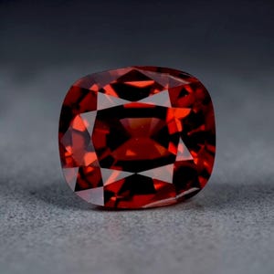 May include: A faceted, cushion-cut, red gemstone. The gem is a deep, rich red color with multiple facets that reflect light. The gemstone is set against a gray background.