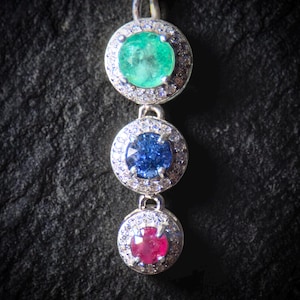 May include: A silver pendant with three circular gemstones: emerald green, sapphire blue, and ruby red. Each gem is encircled by a halo of small, clear stones. The pendant hangs vertically, displaying the vibrant colours of the gems.