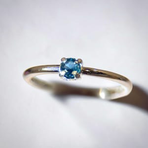 May include: A silver ring with a small, round, blue gemstone. The gemstone is set in a four-prong setting, and the band is thin and polished. The ring is displayed on a white background.