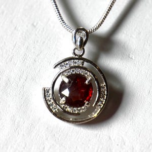 May include: A silver pendant necklace featuring a round, deep red gemstone at the center, surrounded by a halo of small, clear crystals. The pendant hangs from a delicate silver chain, creating a classic and elegant jewelry piece.