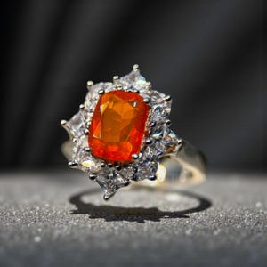 May include: A silver ring featuring a large, rectangular orange gemstone at the center, surrounded by smaller, clear, square-cut stones. The ring's design is set against a dark background, highlighting the sparkle of the gemstones.
