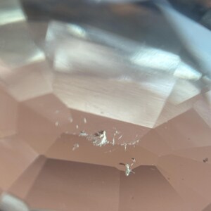 May include: Close-up of a faceted, clear gemstone with a peach hue. The gem's surface reflects light, showcasing its geometric cuts. There are small imperfections visible on the surface. The overall effect is one of clarity and brilliance.