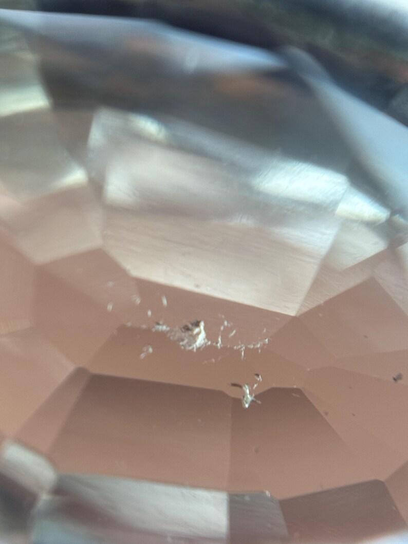May include: Close-up of a faceted, clear gemstone with a peach hue. The gem's surface reflects light, showcasing its geometric cuts. There are small imperfections visible on the surface. The overall effect is one of clarity and brilliance.