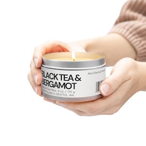Può includere: Una candela rotonda accesa in una latta d'argento è tenuta in entrambe le mani. L'etichetta recita "BLACK TEA & BERGAMOT" e "Cera di soia naturale. 170 g. Hand-Poured in SEATTLE, WA."