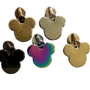Może przedstawiać: Pięć metalowych suwaków w kształcie głowy myszki. Suwaki są w różnych kolorach, w tym złotym, srebrnym, czarnym, tęczowym i mosiężnym. Nadają się do toreb, odzieży i innych projektów krawieckich.