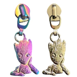 Op de afbeelding: Twee ritsen met een cartoonfiguur. De ene rits is regenboogkleurig en heeft een regenboogkleurige afwerking, de andere is goudkleurig. Beide ritsen hebben een cartoonfiguur met een glimlachend gezicht en een boomvormig lichaam.