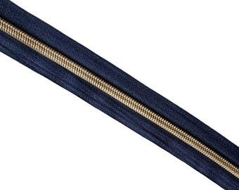 May include: A close-up of a navy blue zipper with a gold-colored metal coil. The zipper is angled diagonally across the frame against a white background. The zipper teeth are tightly interlocked.