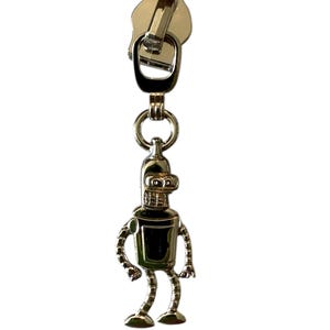 May include: Silver-colored zipper pull featuring a cartoon robot character. The robot has a rectangular body, curved arms, and bent legs. The zipper pull is attached to a metal ring and a larger zipper head.