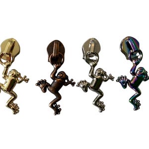 Może przedstawiać: Cztery metalowe suwaki do zamków błyskawicznych z motywami żab. Suwaki są w kolorze złotym, brązowym, srebrnym i opalizującym. Każda żaba trzyma pierścień suwaka. Tło jest białe.