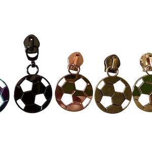 May include: Five soccer ball zipper pulls in various metallic finishes: iridescent, black, copper, bronze, and gold. Each pull features a soccer ball charm with a zipper head attached by a small ring. The soccer balls have a geometric design.