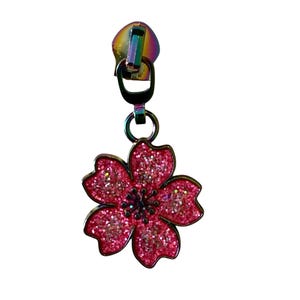 May include: A decorative zipper pull featuring a pink glittery flower charm. The flower has five petals and a dark center. The zipper pull is attached to a rainbow-colored metal zipper head.