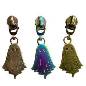 Op de afbeelding: Drie metalen ritstrekkers met spookvormige bedels. Twee hebben een bronzen afwerking en één heeft een regenboogkleurige iriserende afwerking. De spookbedels hebben twee ooggaten en een golvende onderrand.