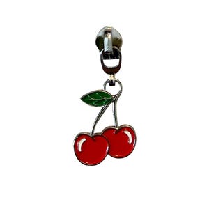 May include: A silver-coloured zip pull with a cherry charm. The charm features two red cherries with a green leaf, all with a glossy finish. The zip pull is attached to a metal ring.