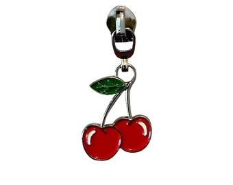 May include: A silver-toned zipper pull with a cherry charm. The charm features two red cherries with a green leaf, all with a glossy finish. The zipper pull is attached to a metal ring.