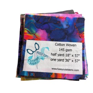 May include: A stack of colorful cotton woven fabric pieces. The fabric has a vibrant abstract design in shades of blue, pink, and orange. A white label displays the text "FUSSY CUTS CUSTOM FABRIC" and fabric details: half yard 18" x 57", one yard 36" x 57".