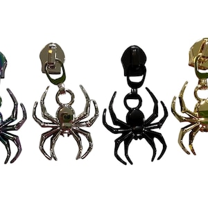 May include: Four metal zipper pulls, each featuring a spider-shaped charm. The set includes one iridescent, one silver-toned, one solid black, and one gold-toned spider charm, each attached to a matching zipper slider component for garment attachment.