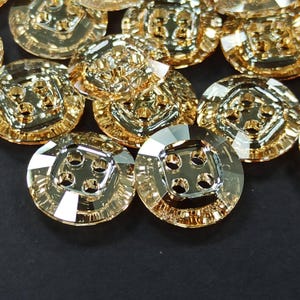 May include: A collection of shiny, gold-colored buttons. Each button has a square center with four holes and a faceted, round outer edge. The buttons are arranged on a dark surface, reflecting light and creating highlights.