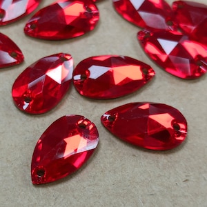Op de afbeelding: Close-up van verschillende gefacetteerde, druppelvormige rode strass-steentjes. Elke edelsteen heeft een gat aan de bovenkant om te rijgen of te naaien. De edelstenen zijn verspreid over een neutrale ondergrond, die hun briljante, reflecterende facetten benadrukt.